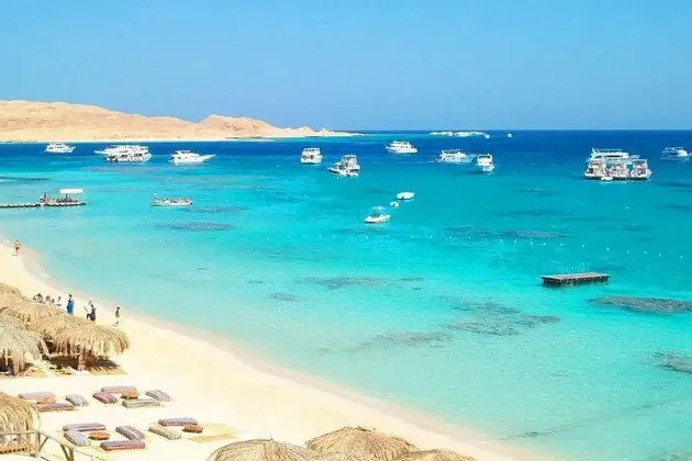 Une plage de sable fin, parsemée de parasols en chaume, surplombe une baie turquoise et limpide où flottent de nombreux bateaux blancs, avec des collines arides en arrière-plan.