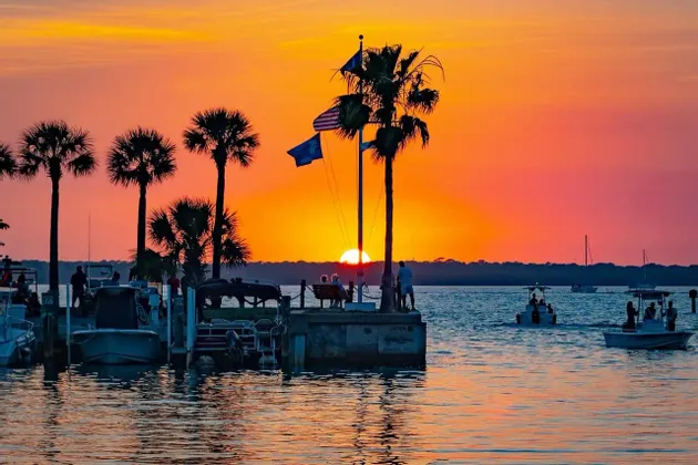 Un tramonto arancione vibrante sull'acqua, con palme e persone in silhouette che osservano da un molo con barche.