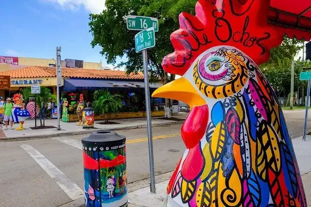 Una grande statua di gallo, dipinta a colori vivaci, si trova su un marciapiede cittadino accanto a un segnale stradale sotto un cielo sereno.