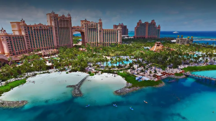 Veduta aerea di un grande resort tropicale con hotel rosa, una spiaggia di sabbia bianca, una laguna turchese e numerose palme.