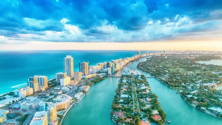 Una veduta aerea di una città costiera con grattacieli lungo l'oceano e isole in un canale turchese sotto un cielo nuvoloso.