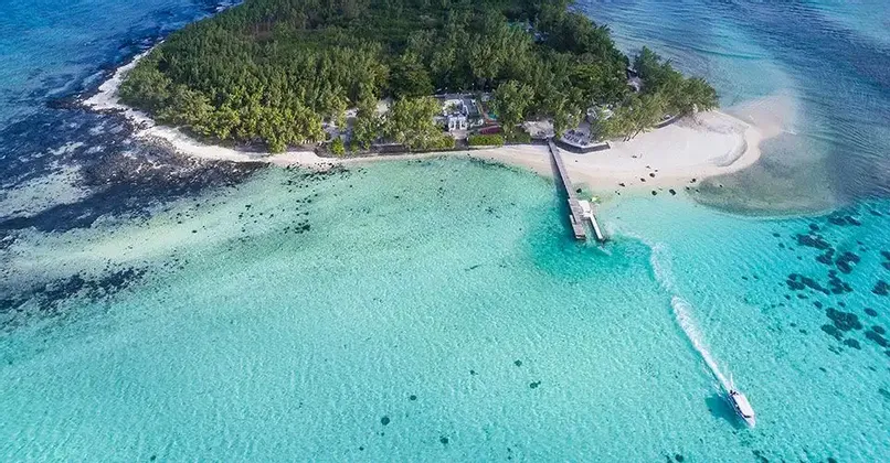 Una vista aerea di una piccola isola ricoperta di alberi, con una spiaggia sabbiosa e un molo, circondata da acqua cristallina turchese con un motoscafo che la attraversa.