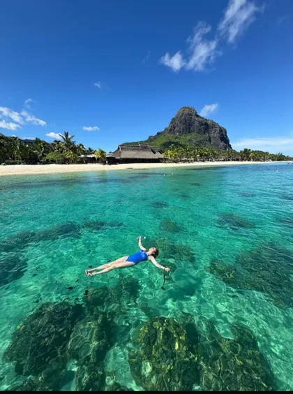 Una mujer en traje de baño azul flota de espaldas en agua clara turquesa cerca de una playa tropical con una montaña al fondo.