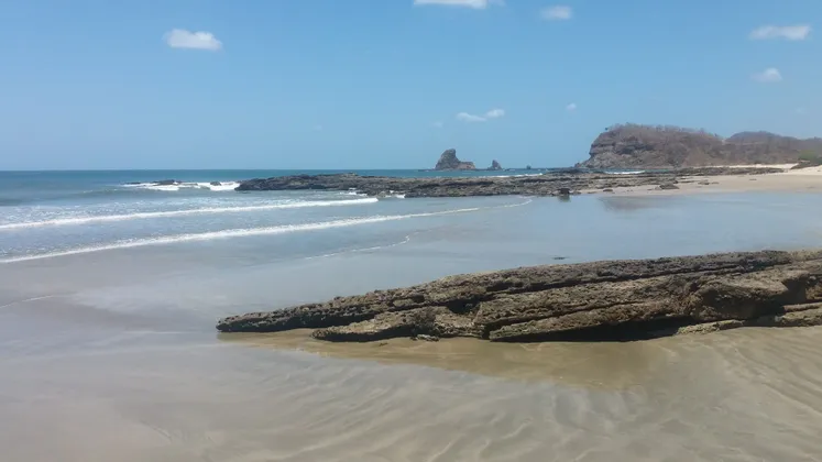 Ein Sandstrand mit großen, dunklen Felsformationen am Ufer und im Meer unter einem klaren blauen Himmel.