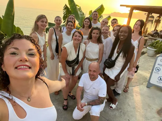 Un groupe WeRoad, tout de blanc vêtu, prend un selfie de groupe au bord de la mer sous un coucher de soleil doré.
