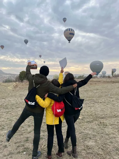 Da dietro, tre persone di un viaggio di gruppo WeRoad si abbracciano e alzano le braccia verso mongolfiere che volano in un cielo nuvoloso.