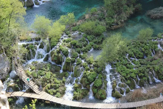 Una vista aerea di persone che camminano su una passerella di legno che si snoda tra cascate terrazzate e piscine turchesi.