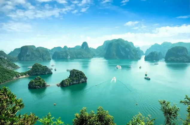 A high-angle view of a turquoise bay with many green limestone islands and several boats sailing on the water under a clear blue sky.