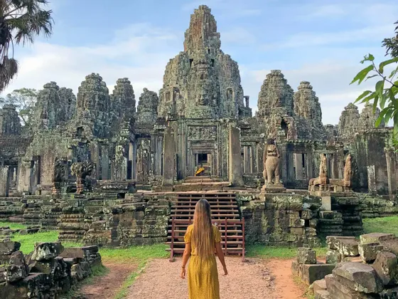 A woman with long hair in a yellow dress walks on a path towards the entrance of a large, ancient stone temple with carved towers.