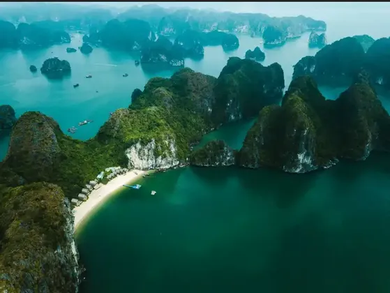 An aerial view of numerous green-topped limestone islands rising from turquoise water, with a secluded sandy beach in the foreground.