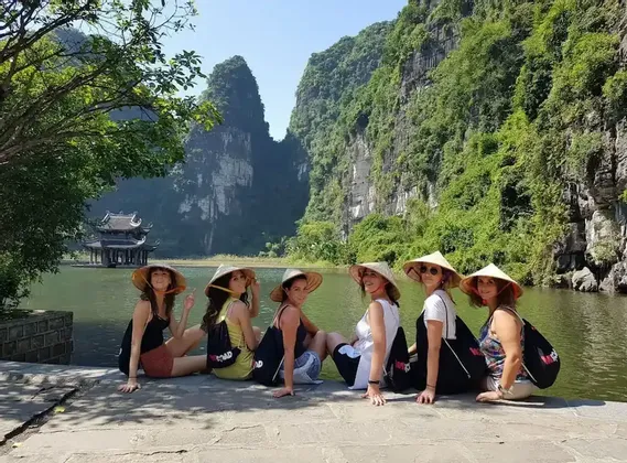 A WeRoad group trip of six women wearing conical hats, sitting by a river with green mountains in the background.