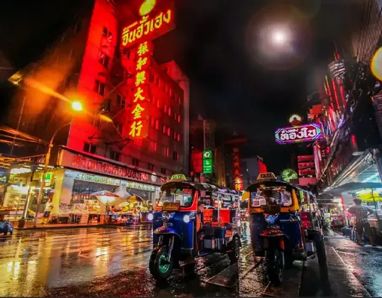 Two tuk-tuks are parked on a wet city street at night, illuminated by bright red and colorful neon signs on buildings.