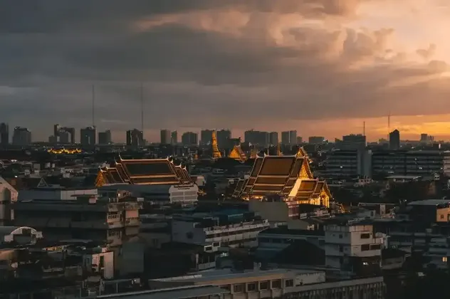 An urban skyline with ornate temple roofs in the foreground, illuminated by a warm sunset under a cloudy sky.