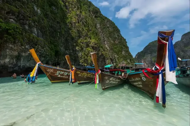 Traditional long-tail boats with colorful ribbons float on clear turquoise water beside a large, green-covered cliff.