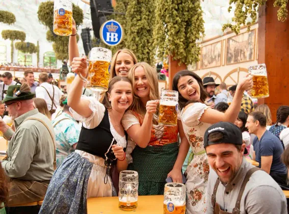 A WeRoad group trip of young people in traditional attire smiling and raising their beer mugs in a festive beer hall.