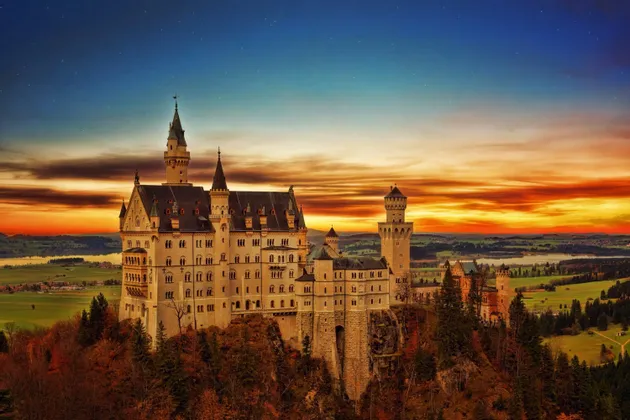A large castle with multiple towers sits on a forested cliff, overlooking a valley with lakes under a colorful sunset sky.