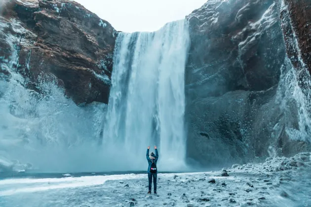 Une personne, les bras levés, fait face à une immense cascade dévalant des falaises sur un rivage glacé.