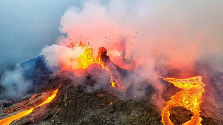 Ein aktiver Vulkan bricht aus, geschmolzene Lava strömt in Flüssen seine dunkle, felsige Flanke hinab unter einem rauchgeschwängerten Himmel.