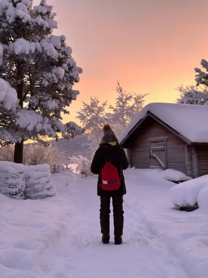 Eine Person mit rotem WeRoad-Rucksack steht bei Sonnenuntergang auf einem verschneiten Weg neben einer Holzhütte.