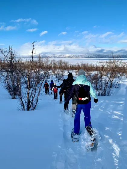 Eine WeRoad-Gruppe wandert im Gänsemarsch mit Schneeschuhen durch eine verschneite Landschaft mit kahlen Bäumen, auf einen zugefrorenen See und Berge zu.