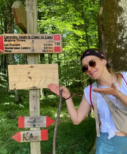 Eine lächelnde Frau mit Sonnenbrille und Kopftuch zeigt auf einen hölzernen Wanderwegweiser in einem üppig grünen Wald.