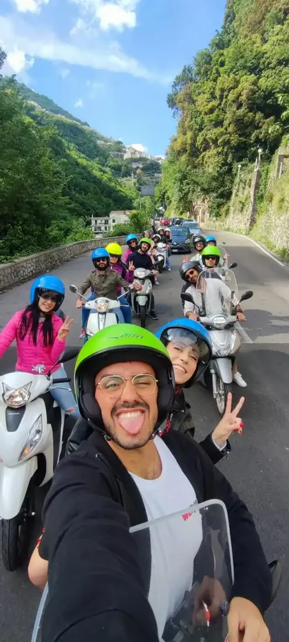 Un gruppo WeRoad in viaggio su scooter si scatta un selfie mentre percorre in fila una strada tortuosa accanto a una collina verde.