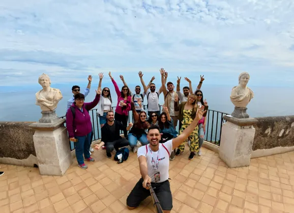 Un gruppo WeRoad in viaggio scatta un selfie su una terrazza panoramica con busti in marmo, in posa e sorridendo con il mare sullo sfondo.