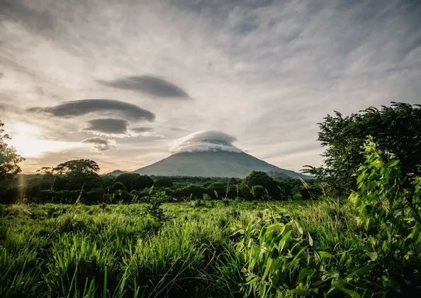 Un volcan dont le sommet est couvert par un nuage lenticulaire, vu depuis un champ herbeux sous un ciel nuageux au coucher du soleil.