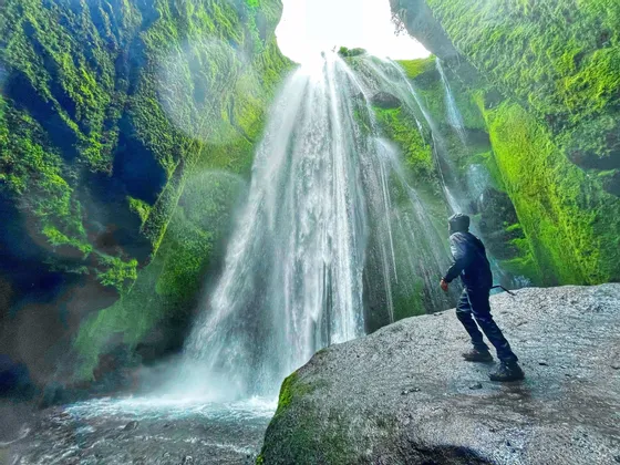 Eine Person steht auf einem nassen Felsen und blickt zu einem Wasserfall auf, der die grünen, moosbewachsenen Wände einer Schlucht herabstürzt.