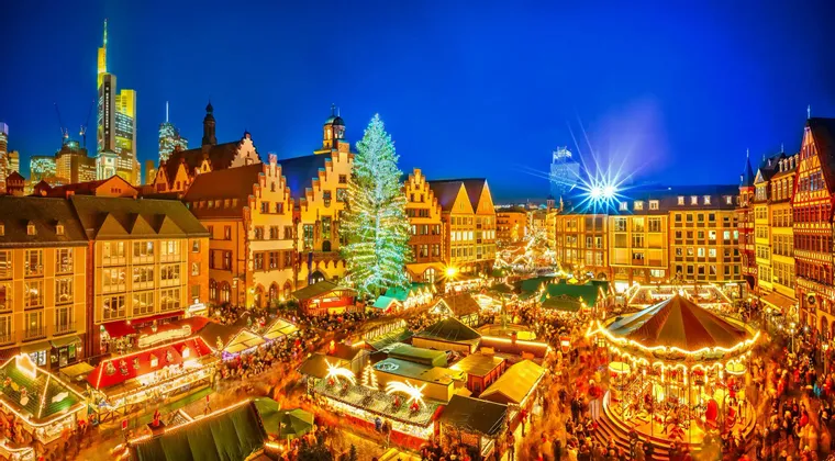 An aerial view of a crowded Christmas market at night, with an illuminated tree, a carousel, and historic buildings set against a modern city skyline.