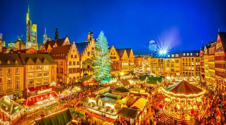 An elevated view of a bustling Christmas market in a city square at night, with a large illuminated tree, a carousel, and festive stalls.