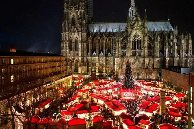 An elevated view of a bustling Christmas market at night, with red-roofed stalls and a large tree, set against an illuminated Gothic cathedral.