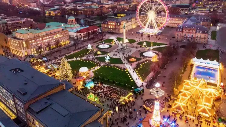 An aerial view of a bustling city square at dusk, filled with a Christmas market, an illuminated Ferris wheel, and an ice rink.