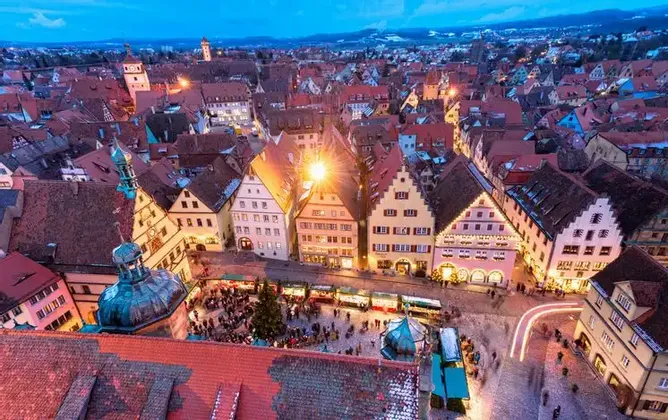 An aerial view of a historic European town square bustling with a Christmas market at twilight, with lights illuminating the gabled buildings.