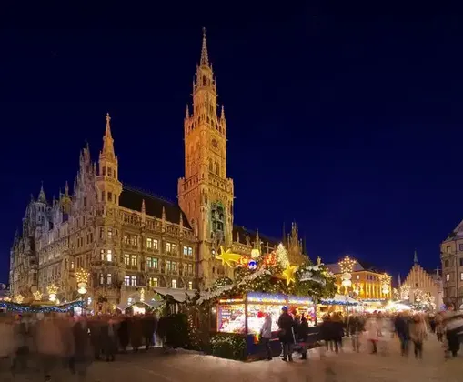 A bustling Christmas market at night, with motion-blurred crowds and festive stalls in front of an illuminated Gothic town hall.
