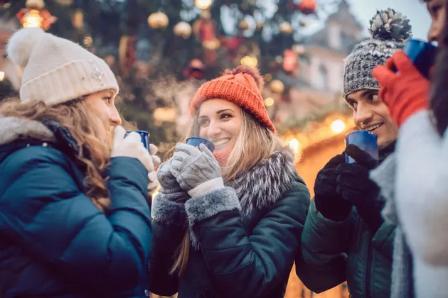 A WeRoad group trip of friends in winter hats and coats, smiling and drinking hot beverages from mugs at an outdoor market.