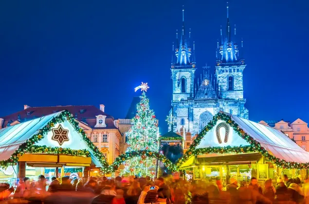 A crowded Christmas market at night with an illuminated tree and stalls in front of a large Gothic church.