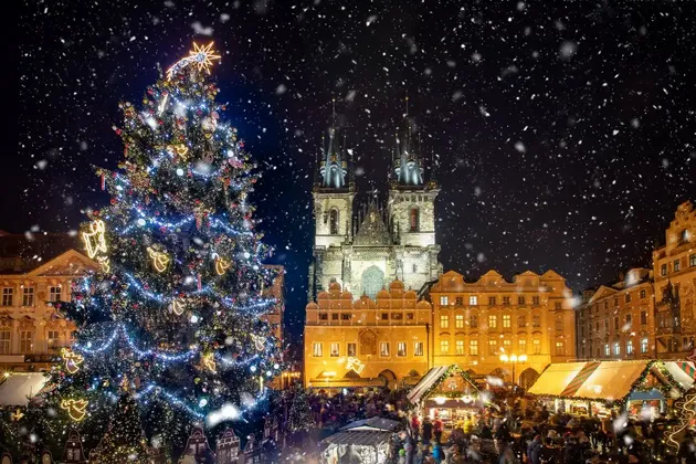 A large, illuminated Christmas tree stands in a bustling town square during a snowy Christmas market at night.