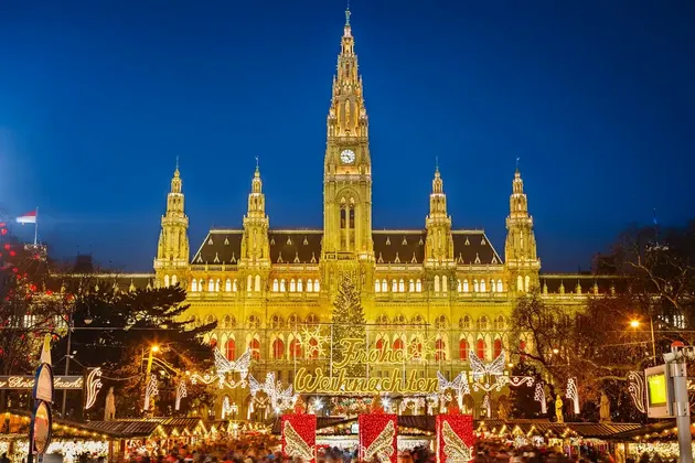 A large, illuminated building with a clock tower at night behind a bustling Christmas market with festive lights and a tree.