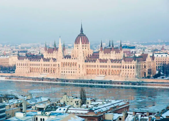 A grand, ornate building with a central dome sits on the banks of an icy river, with snow-covered city rooftops in the foreground.