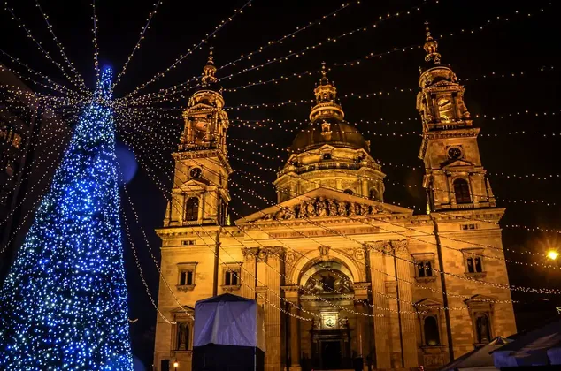An illuminated basilica at night, with a large Christmas tree covered in blue lights and festive string lights overhead.