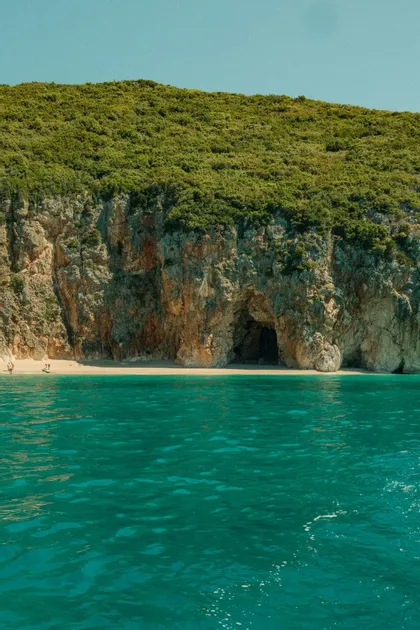 Una vista desde el mar turquesa de una playa de arena aislada con una cueva al pie de un acantilado rocoso y frondoso.
