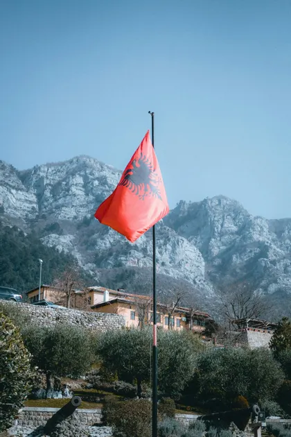 La bandera albanesa ondea en un mástil frente a una gran cordillera rocosa bajo un cielo despejado.