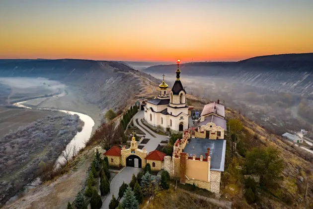 Una vista aerea di un monastero con cupole dorate arroccato su una scogliera che domina un fiume serpeggiante in una valle nebbiosa all'alba.