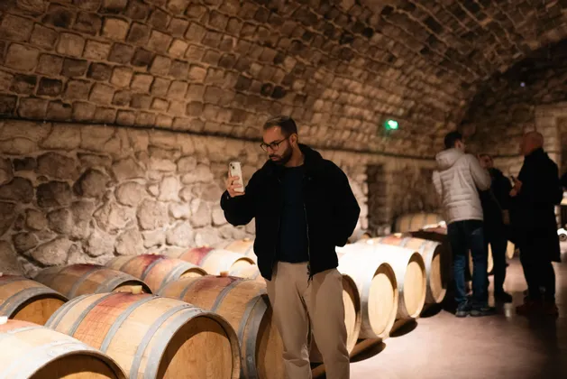 Un uomo in viaggio di gruppo WeRoad usa lo smartphone per fotografare botti di vino in legno in una cantina di pietra.