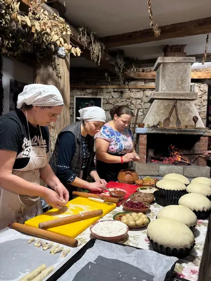 Tre donne preparano impasto e dolci su un lungo tavolo in una cucina rustica con un grande camino in pietra.