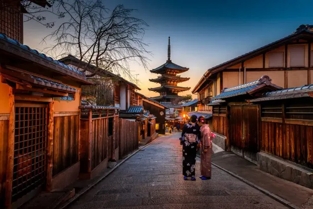 Due donne in kimono tradizionali su una strada acciottolata osservano un'alta pagoda al tramonto.