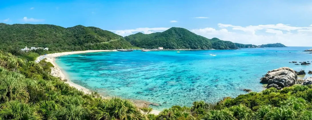 Vista panoramica di una baia tropicale con spiaggia sabbiosa, acqua turchese cristallina e rigogliose colline verdi sotto un cielo azzurro.