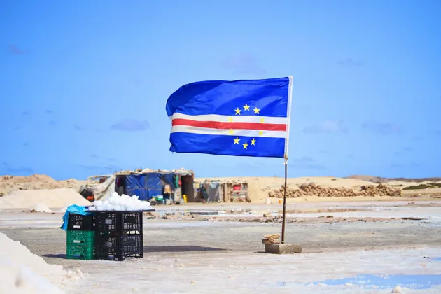 La bandera de Cabo Verde ondea al viento sobre un vasto salar con pequeños edificios en la distancia bajo un cielo azul.