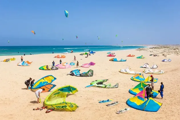 Una spiaggia soleggiata piena di gente che prepara colorate attrezzature da kitesurf, con molti aquiloni già in volo sull'oceano.
